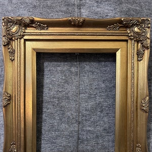 May include: Ornate gold picture frame with detailed corner and edge embellishments. The frame has a rectangular opening and a slightly curved outer edge. The gold finish gives the frame a vintage or antique appearance.