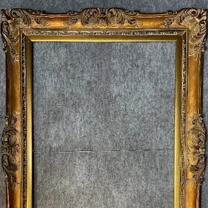 May include: A rectangular picture frame with a metallic gold finish. The frame features ornate, raised relief carvings along the edges, including scrollwork and floral motifs at the corners and centers of each side, surrounding a plain inner border.
