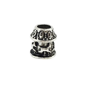 May include: Silver-toned metal bead with intricate designs. The bead has a cylindrical shape with a wider top and bottom. The top and bottom of the bead are decorated with a circular pattern of raised dots and ovals. The middle section of the bead features a detailed design of interlocking shapes.
