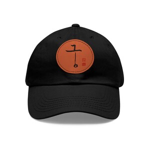 Puede incluir: Gorra de béisbol negra con un parche redondo naranja que presenta un carácter kanji negro sobre fondo blanco. El carácter kanji está rodeado por un cuadrado rojo.