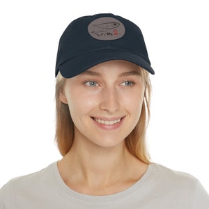 Può includere: Un cappello da baseball blu navy con una toppa marrone che presenta un'illustrazione in bianco e nero di pesci e il testo "Fish Soup".