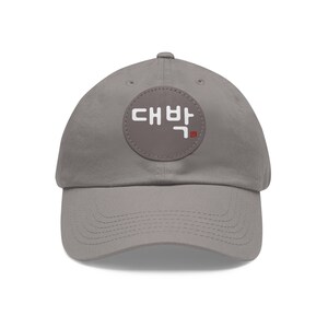 May include: A gray baseball cap with a curved brim. The cap features a circular patch with white Korean text and a small red stamp. The hat is made of a soft fabric and has an adjustable strap.