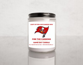 May include: A white candle with a black lid and a red and white Buccaneers logo. The label reads "Light up for a Buccaneers win! Fire the cannons Game Day Candle".