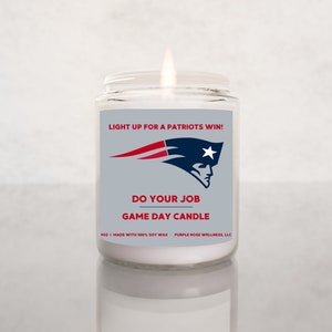 May include: A lit glass jar candle with a label featuring the text "LIGHT UP FOR A PATRIOTS WIN!" and "DO YOUR JOB GAME DAY CANDLE". The label also displays a logo with a red, white, and blue design. The candle is 9 oz and made with 100% soy wax.