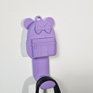 Puede incluir: Un gancho de pared con forma de mochila de color lavanda con un lazo y orejas. Una bolsa negra cuelga del gancho.