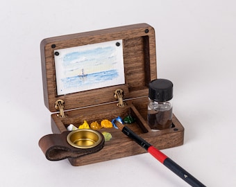 Puede incluir: Un juego de acuarela de viaje de madera con un caballete con bisagras, una pequeña paleta de pintura, una botella de agua y un pincel. El juego está abierto y el caballete está levantado.