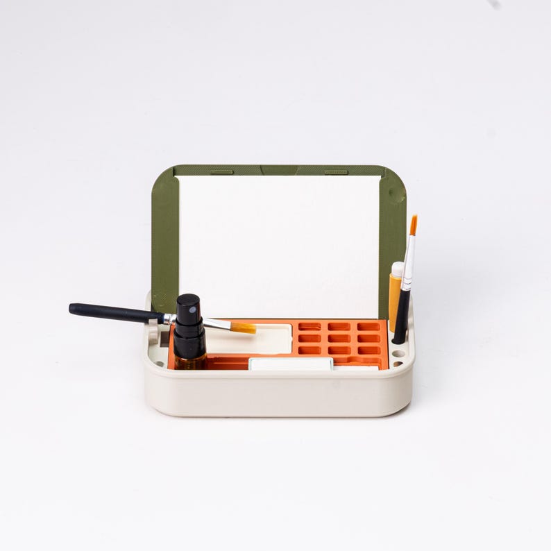 May include: A beige and olive green artist's palette box with an open lid. The box contains a black spray bottle, paintbrushes, and orange compartments for paint. The box is designed for art supplies.