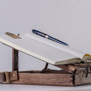 Puede incluir: Un escritorio de madera con una correa de cuero, un cuaderno y un bol&iacute;grafo azul. El escritorio est&aacute; abierto y el cuaderno est&aacute; apoyado en &aacute;ngulo. El escritorio est&aacute; hecho de madera oscura y tiene un acabado natural.