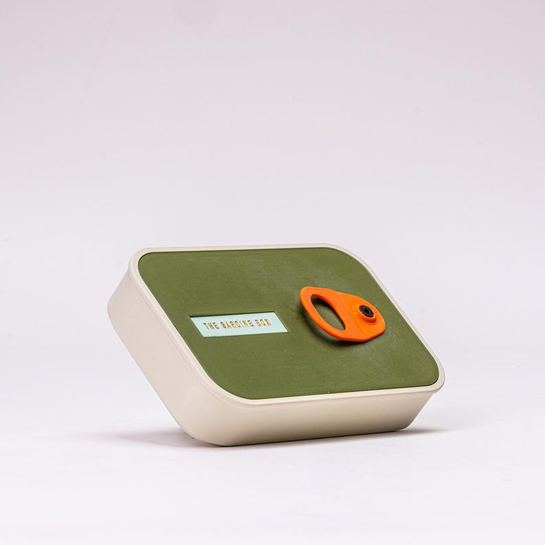 May include: A rectangular container with a rounded top and base. The top is olive green with an orange handle and the text "THE BARDING BOX". The base is off-white.