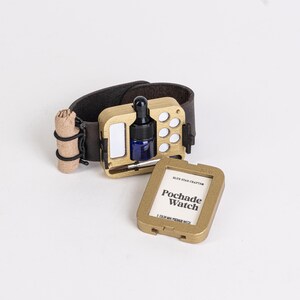 May include: A gold-colored Pochade Watch with a dark brown leather wristband. The watch features a small blue bottle with a dropper, a mirror, and a compartment for paint. The watch also includes a small, rolled-up item and a separate gold-colored case with the text "Pochade Watch."