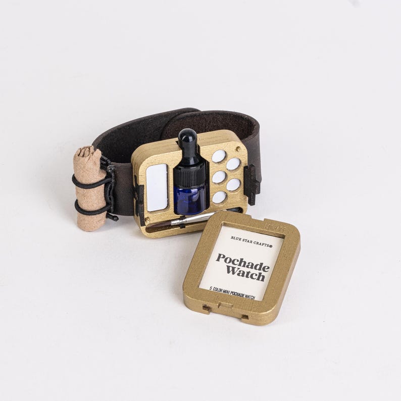 May include: A gold-colored Pochade Watch with a dark brown leather wristband. The watch features a small blue bottle with a dropper, a mirror, and a compartment for paint. The watch also includes a small, rolled-up item and a separate gold-colored case with the text "Pochade Watch."