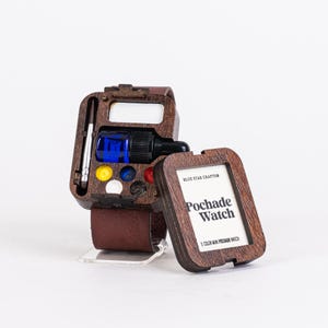Puede incluir: Un Pochade Watch de madera con una correa de cuero marrón. La caja del reloj contiene una botella azul, un pincel y varios colores de pintura. La esfera del reloj dice "Pochade Watch" y "5 Color Mini Pochade Watch".