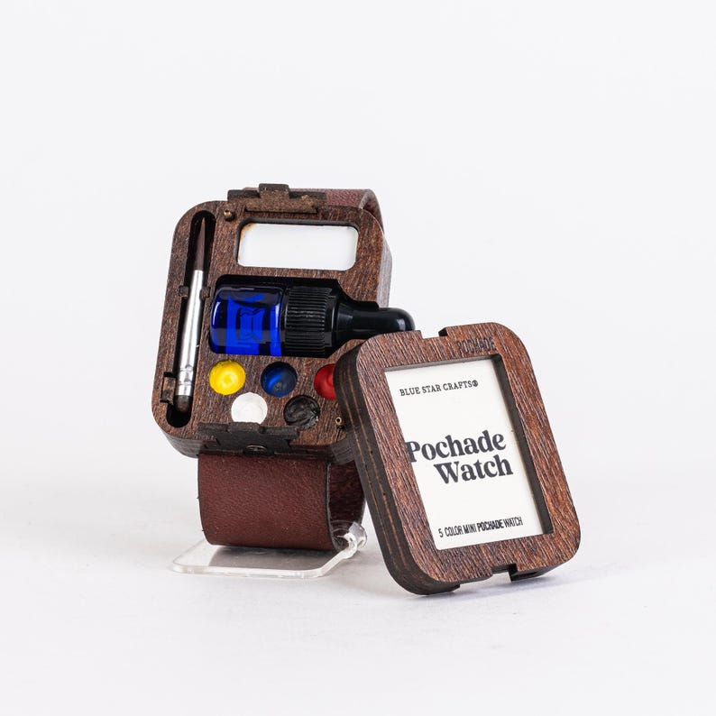 May include: A wooden Pochade Watch with a brown leather strap. The watch case contains a blue bottle, a brush, and several paint colors. The watch face reads "Pochade Watch" and "5 Color Mini Pochade Watch".