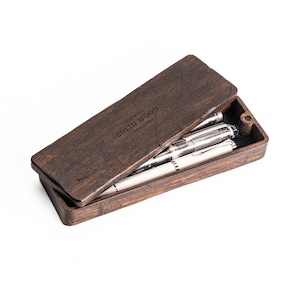 Puede incluir: Un estuche de madera marrón oscuro para bolígrafos con las palabras "Copper Wood Solid Wood Paris, NY 1974" grabadas en la tapa. El estuche está abierto y contiene tres bolígrafos plateados.