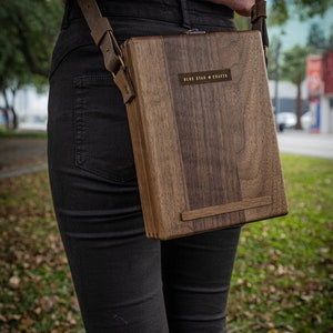 Puede incluir: Una persona que lleva pantalones vaqueros negros y una camisa rosa lleva un bolso de bandolera de madera con una correa de cuero marr&oacute;n. El bolso tiene un cierre de lat&oacute;n y una placa de lat&oacute;n con el texto "BLUE STAR CRAFTS" en &eacute;l.