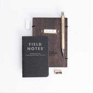 Puede incluir: Un cuaderno negro Field Notes con una etiqueta que dice "48-Page Memo Book", una cubierta de madera con una etiqueta de latón, un lápiz mecánico dorado y un sacapuntas dorado.