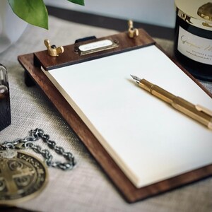 May include: A wooden clipboard with a blank white notepad and a gold pen. The clipboard has a brass latch and a label that says "Notepad".