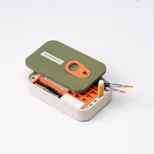 May include: An open, rectangular art supply box with a green lid and orange accents. The lid has the text "THE BARNING BOX" and an orange handle. Inside are paintbrushes, a pencil, and other art supplies. The box is beige.