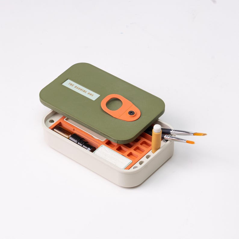 May include: An open, rectangular art supply box with a green lid and orange accents. The lid has the text "THE BARNING BOX" and an orange handle. Inside are paintbrushes, a pencil, and other art supplies. The box is beige.