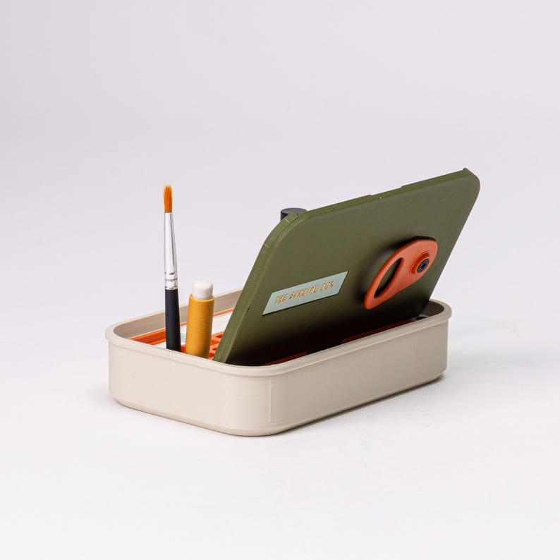 May include: A beige rectangular art supply organizer with an olive green lid. Inside are a paintbrush, a small tube, and the text "THE POTTER'S CO." The lid has an orange handle.