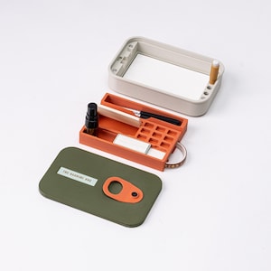 May include: An open, rectangular, multi-compartment case in shades of orange, gray, and olive green. The case contains a small bottle, a pen, and other items. The lid is olive green with an orange cutout. The text "THE BARBIRE BOX" is visible on the lid.