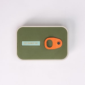 May include: A rectangular olive-green box with a light blue label that reads "THE SARDINE BOX." An orange, circular pull-tab is on the right side. The box has a silver-colored border.