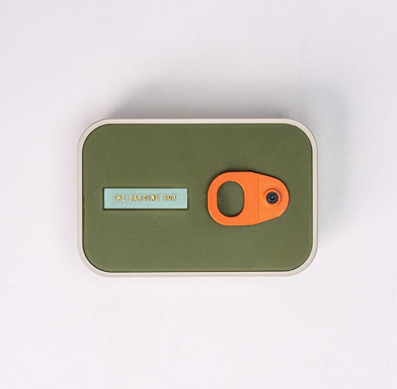 May include: A rectangular olive-green box with a light blue label that reads "THE SARDINE BOX." An orange, circular pull-tab is on the right side. The box has a silver-colored border.