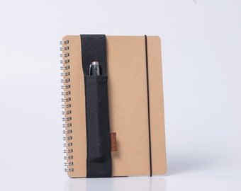 Peut inclure: Un carnet brun avec un élastique noir et un porte-stylo. Le carnet a une reliure spirale et un stylo noir est inséré dans le porte-stylo.