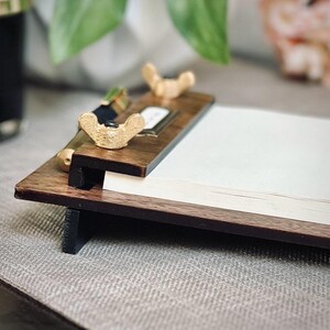 May include: A wooden clipboard with a notepad and two gold metal clips. The clipboard has a dark brown finish and a black base.