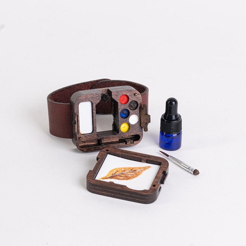 May include: A wooden watercolor paint set with a brown leather wrist strap. The set includes a small blue bottle, a paintbrush, and a small painting of a leaf. The paint set has red, blue, yellow, and white paint colors.