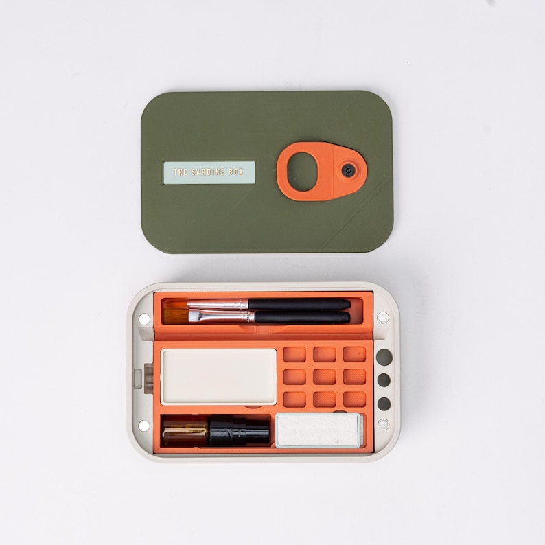 May include: An open, olive green and orange makeup organizer with a light beige frame. The organizer contains makeup brushes, a small mirror, and other beauty supplies. The lid has the text "THE SARDINES BOX".