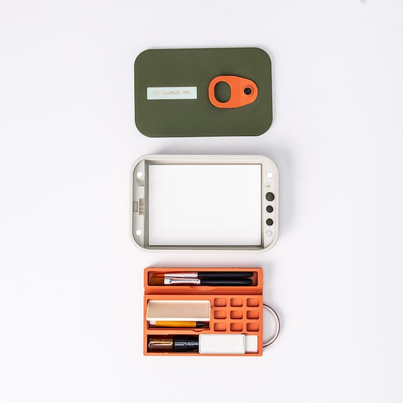 May include: An olive green case with an orange handle and a white label. Below it is a white rectangular frame. At the bottom is an orange pencil case with various pens, pencils, and erasers. The pencil case has a silver ring.