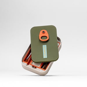 May include: An open, beige makeup case with an olive green lid. The lid has an orange clasp and a small, rectangular label that reads "THE SAPLINGS CO." Inside, orange compartments hold makeup brushes. The case is designed to hold makeup.