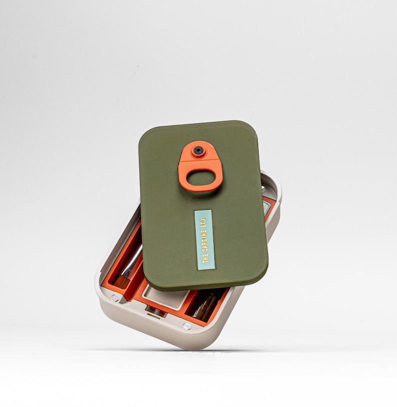 May include: An open, beige makeup case with an olive green lid. The lid has an orange clasp and a small, rectangular label that reads "THE SAPLINGS CO." Inside, orange compartments hold makeup brushes. The case is designed to hold makeup.