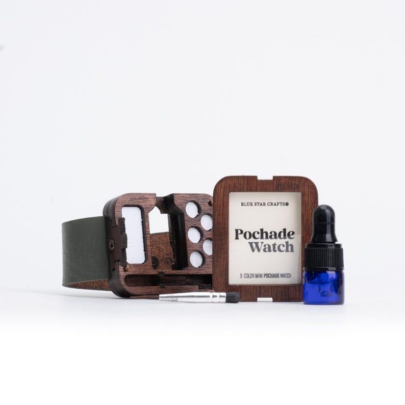 May include: A wooden watch case with a dark green leather strap, a small blue bottle, and a frame with the text "Pochade Watch". The watch case is open, revealing white inserts and circular holes.