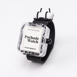 May include: A black wristwatch with a clear, square watch face. The watch face displays the text "Pochade Watch" and "5 Color Mini Pochade Watch". The watch has a black leather strap and a white, cylindrical accessory.