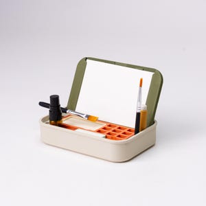 May include: A beige art supply box with an olive green lid, holding a white paper, paintbrushes, and a small bottle. The box has an orange paint palette and a small tube of paint. The art supplies are neatly arranged.