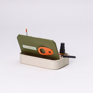 May include: A beige rectangular container with an olive green lid, featuring an orange clasp. The lid is slightly open, revealing a small bottle, a paintbrush, and a black pen. The text "THE WANDER BOX" is visible on the lid.