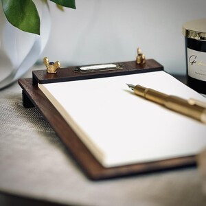 May include: A wooden writing pad with a brass pen holder and a brass label that says "Dear Thoughts". A gold fountain pen is lying on the pad.