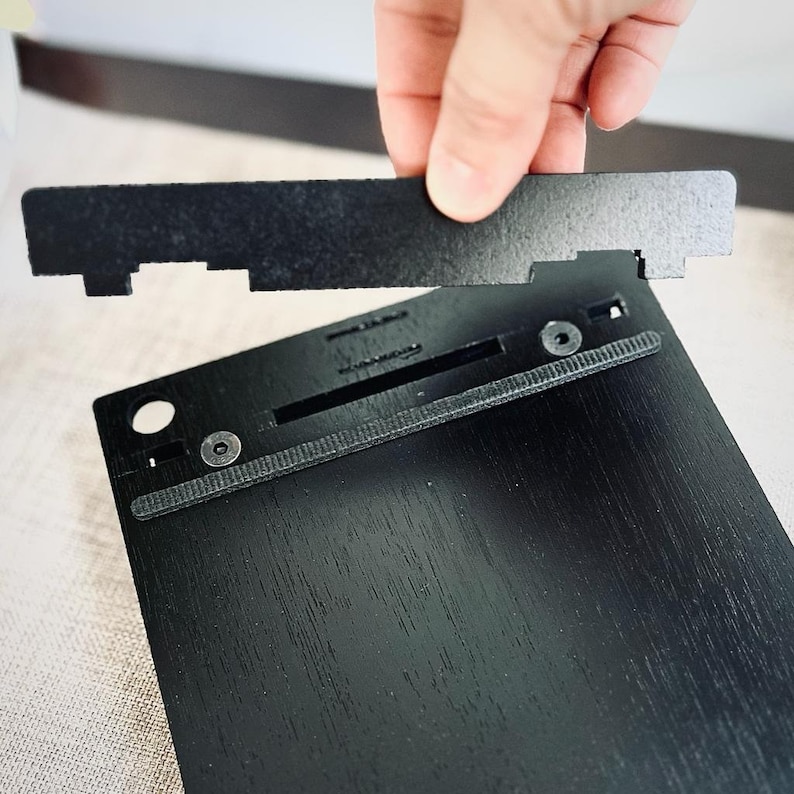 May include: A black wooden frame with a black fabric backing and a black metal latch. The frame is being held by a hand.