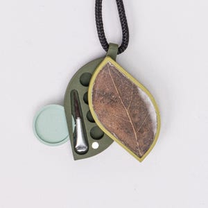 Puede incluir: Un collar con colgante único con una hoja preservada en un marco dorado. Incluye un estuche verde oliva con una pequeña herramienta y un elemento circular azul claro, suspendido de un cordón negro. Diseño inspirado en la naturaleza.