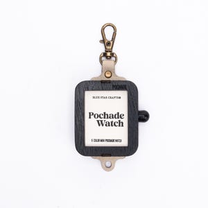 May include: A black wooden Pochade Watch holder with a brass-colored clip. The holder has a rectangular shape with rounded corners and a small black knob. The text "Pochade Watch" is printed on the white center.