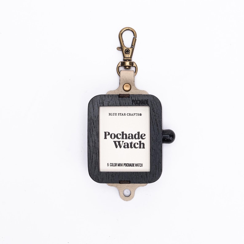 May include: A black wooden Pochade Watch holder with a brass-colored clip. The holder has a rectangular shape with rounded corners and a small black knob. The text "Pochade Watch" is printed on the white center.