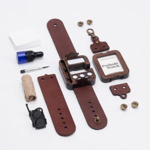 May include: A disassembled wooden watch with a brown leather strap, a small frame with the text "Pochade Watch", and various components like screws, a brush, and a blue bottle. The watch components are arranged on a white surface.