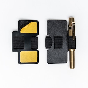 Puede incluir: Dos porta tarjetas de cuero negro con bandas elásticas y cinta adhesiva amarilla. Un bolígrafo dorado con un clip negro está unido al porta tarjetas de la derecha.