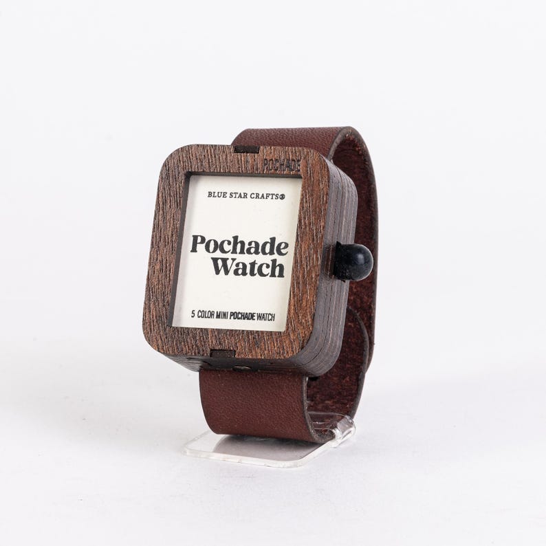 May include: A square-faced wooden watch with a brown leather band. The watch face displays the text "Pochade Watch" and "5 Color Mini Pochade Watch". The watch is displayed on a clear stand.