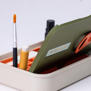 May include: A beige and orange organizer box with a hinged olive green lid. Inside are a paintbrush, a lip balm, and a black bottle. The lid has a label that reads "THE SARDINE BOX".