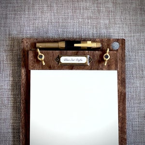May include: A wooden clipboard with a brass pen holder and a brass label that reads "Blue Star Crafts". The clipboard has a white sheet of paper.