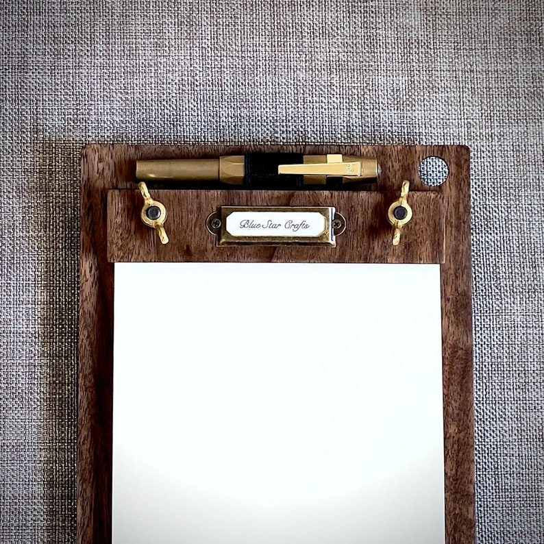 May include: A wooden clipboard with a brass pen holder and a brass label that reads "Blue Star Crafts". The clipboard has a white sheet of paper.