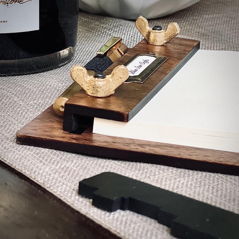 May include: A wooden notepad holder with a brass pen holder and two brass wing nuts. The notepad has the text "Blue Star Coffee" printed on it.